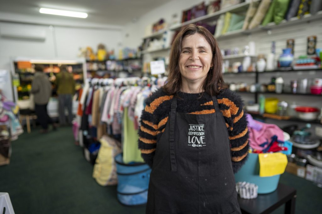 A woman with dark hair, a tiger striped, and an ADRA apron stands in front of a clothing rack in an Op Shop